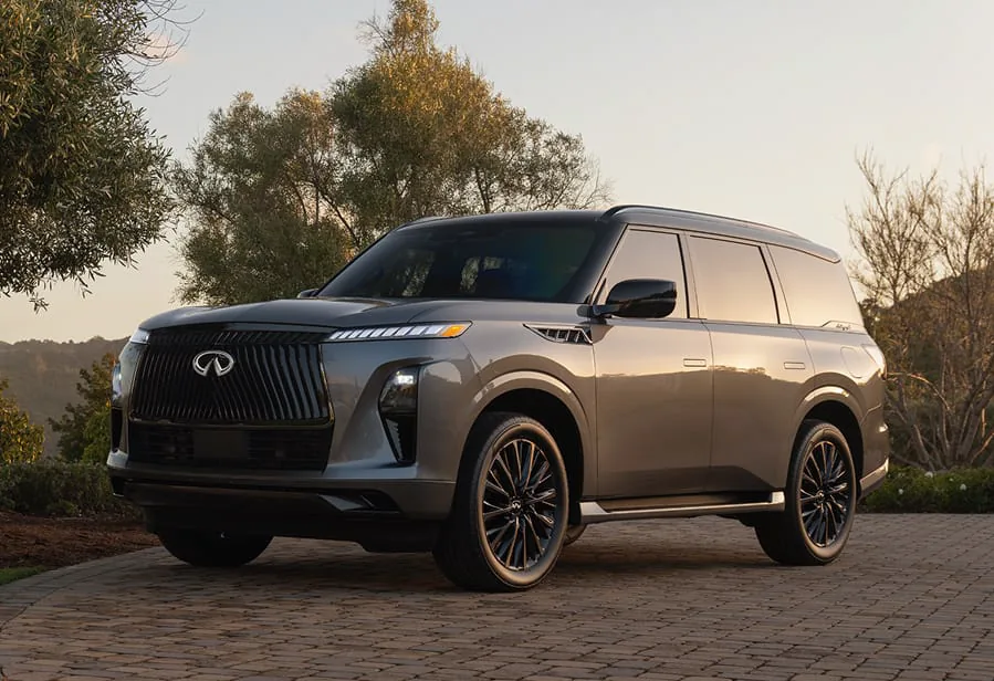 Front three-quarter view of a champagne metallic 2027 INFINITI QX80 with a black roof, parked on cobblestone at golden hour beside olive trees.