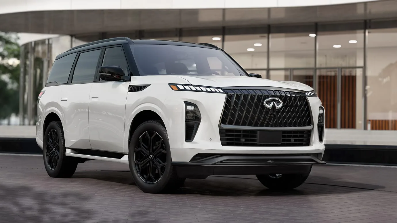 Front three-quarter view of a white 2027 INFINITI QX80 SPORT with a black roof and gloss-black wheels, parked before a modern glass building.