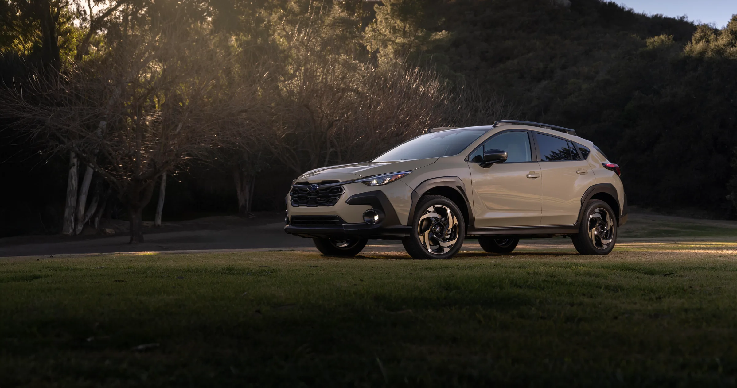 A tan 2027 Subaru Crosstrek Hybrid compact SUV parked on a grassy outcrop at dusk.