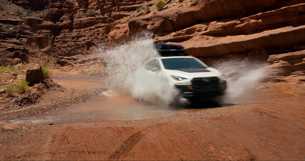 A 2027 Crosstrek Wilderness off-road compact SUV splashes through a puddle in a canyon.