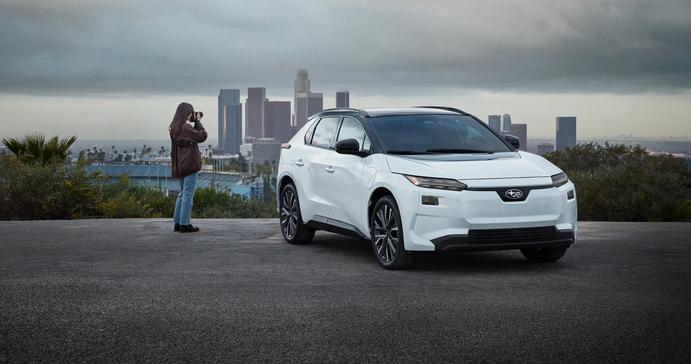 A person takes a photo next to a white Solterra EV parked in front of a city skyline.
