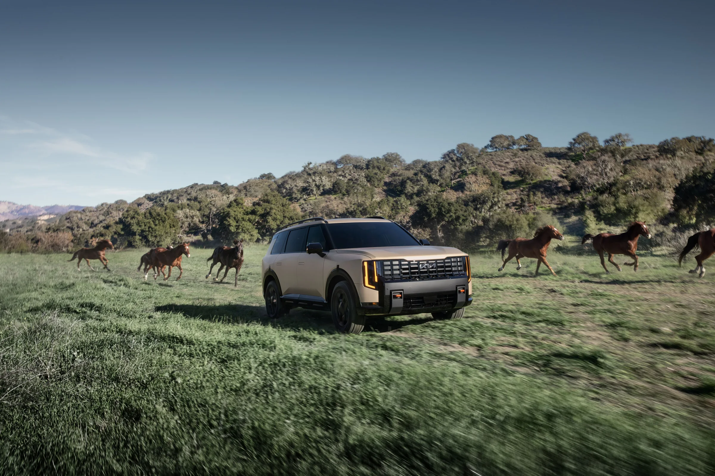 Tan 2027 Kia Telluride X-Pro parked in a grassy field with horses running on hills behind it