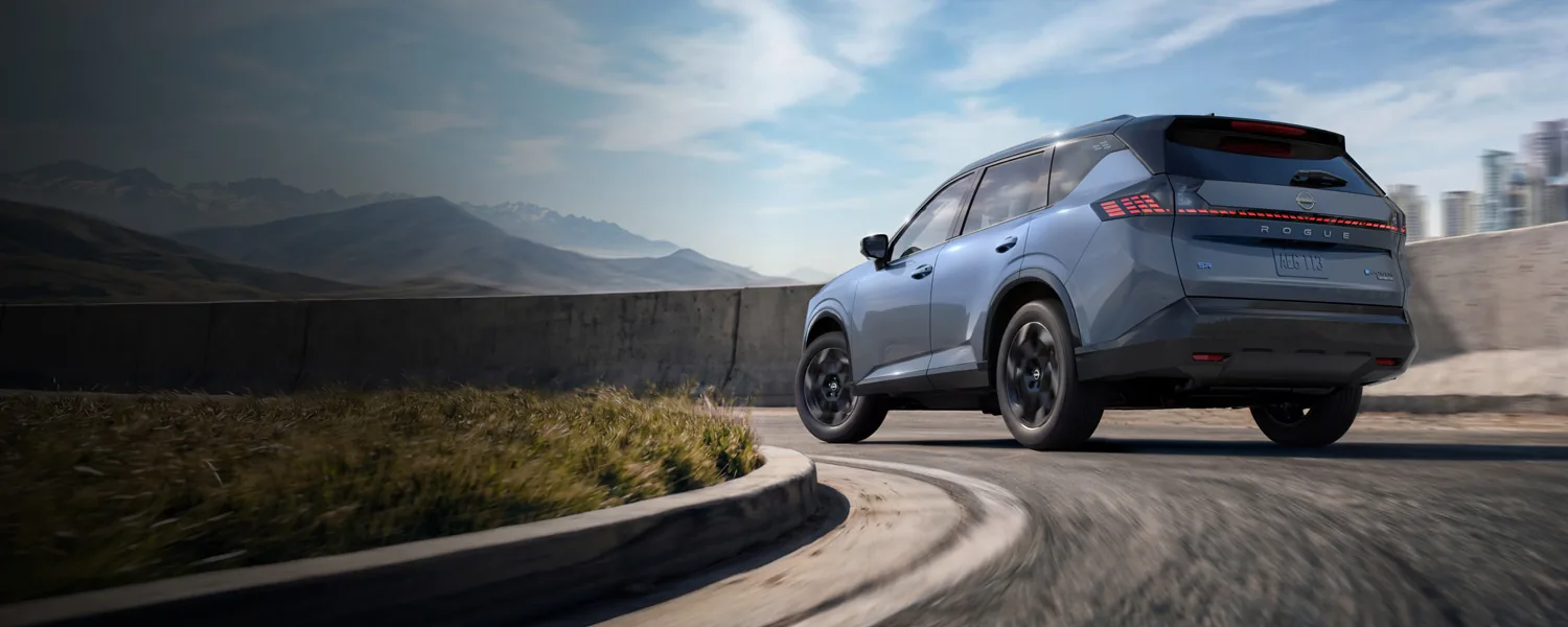 Rear three-quarter view of the 2027 Nissan Rogue Hybrid driving through a curve on an open road.
