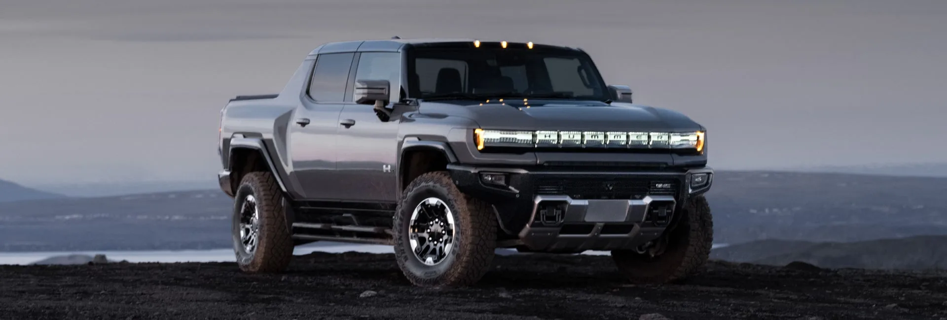 Front three-quarter view of a white 2025 GMC HUMMER EV Pickup driving on a wet city street at dusk with illuminated headlights