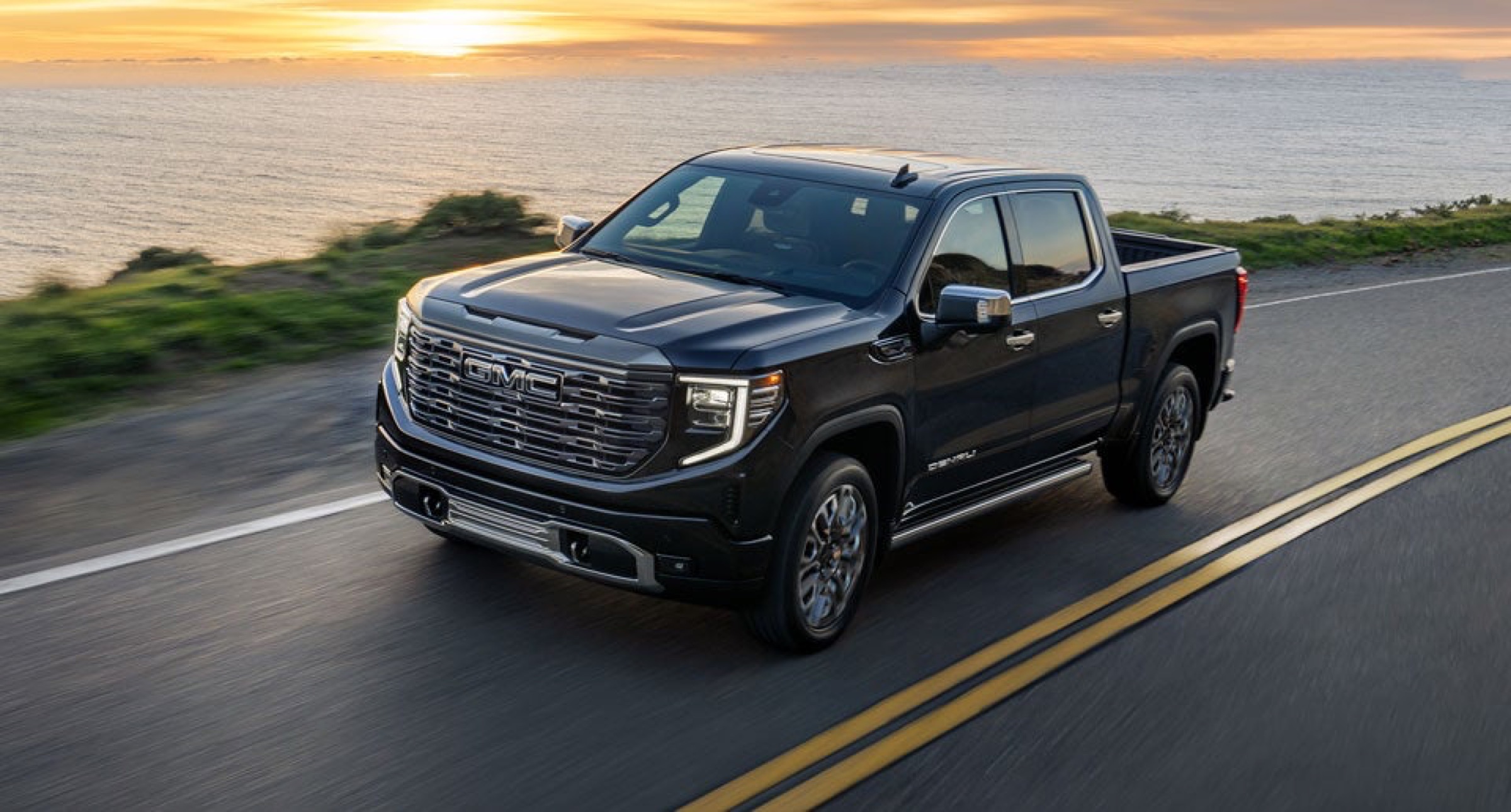 Black GMC Sierra pickup driving on a coastal road at sunset
