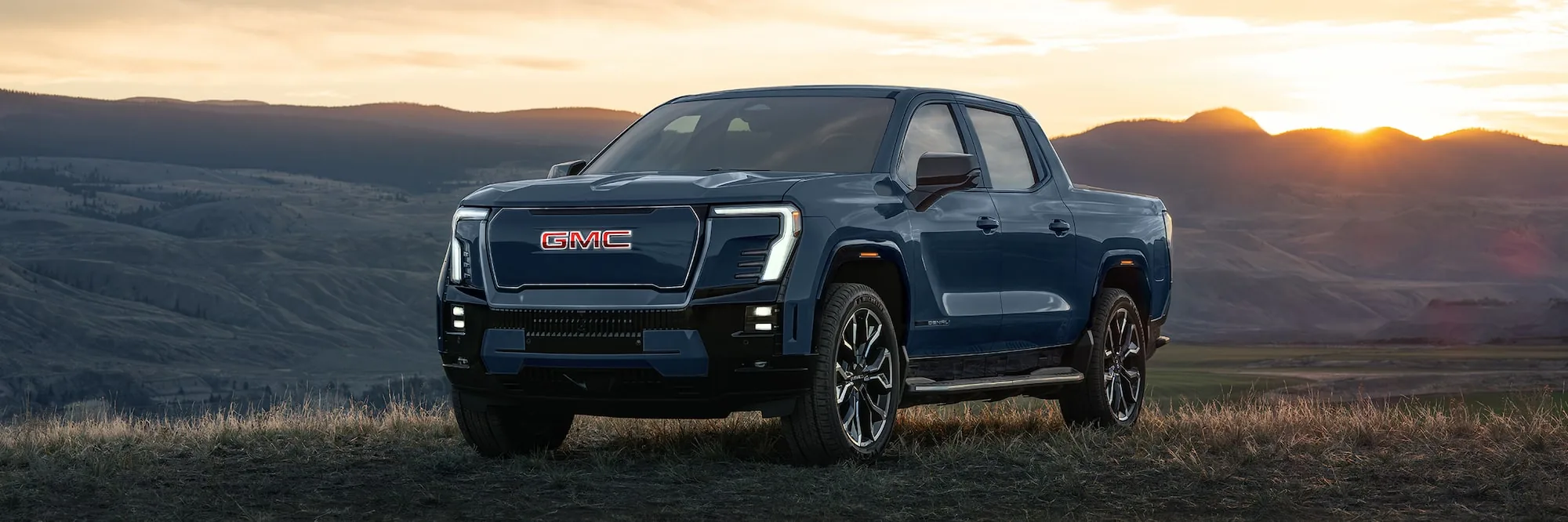 Front three-quarter view of a dark 2026 GMC Sierra EV pickup parked in a valley at sunset with rolling hills in the distance