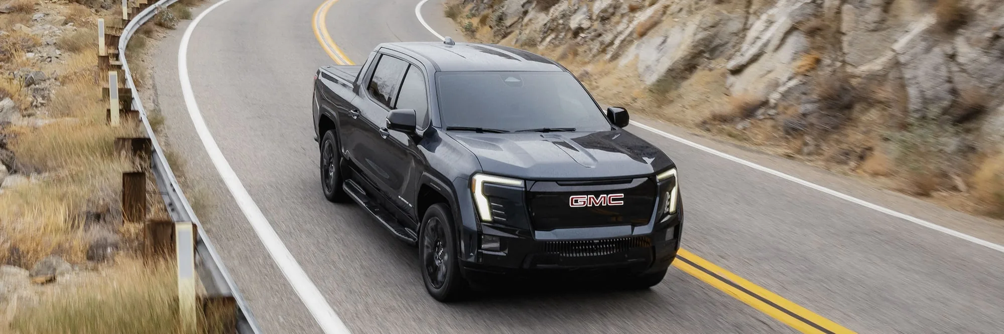 Front three-quarter view of a dark 2026 GMC Sierra EV pickup truck parked in a valley at sunset
