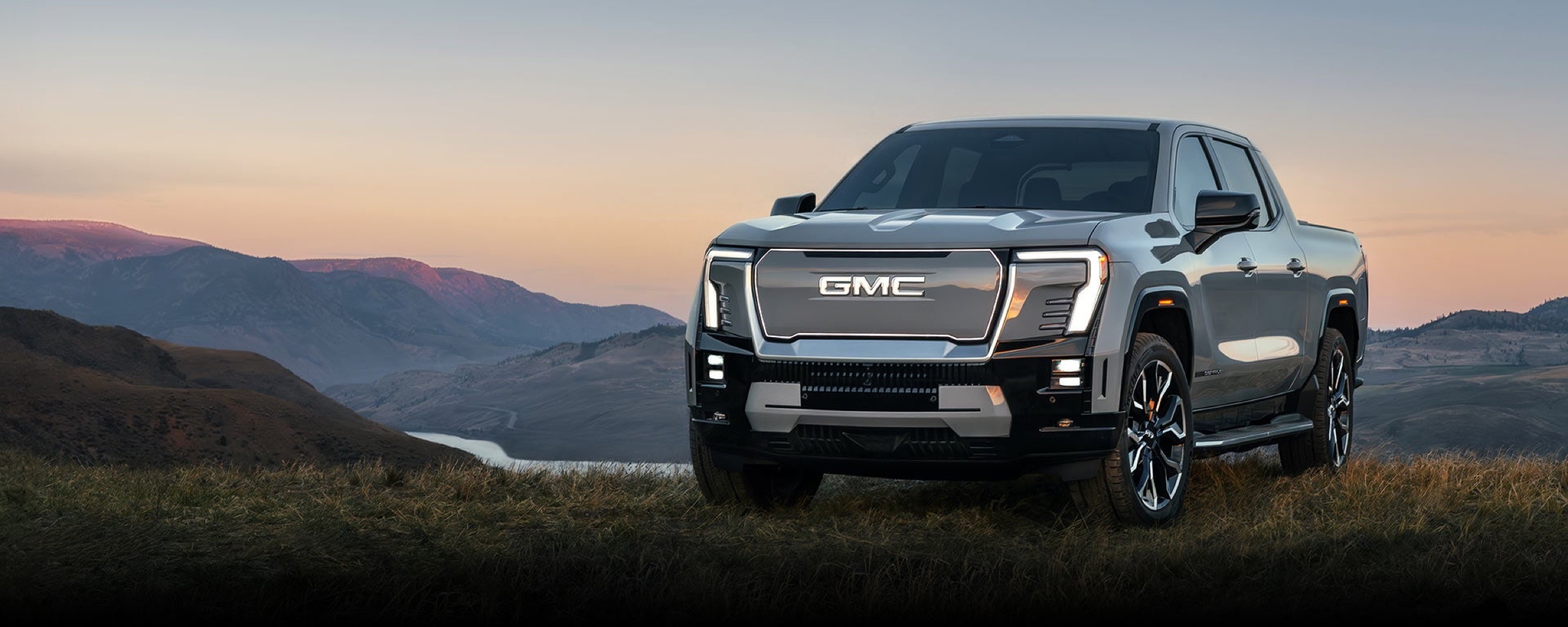 Silver GMC Sierra EV Denali electric pickup parked on a grassy hillside overlooking mountains at dusk